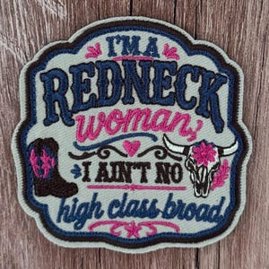 May include: An embroidered patch with a light gray background featuring the dark blue text I'M A REDNECK, pink text woman, dark brown text I AIN'T NO, and dark blue text high class broad. It includes a brown cowboy boot and a white cow skull.