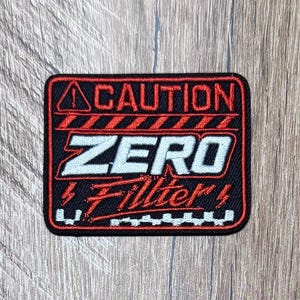 May include: A rectangular embroidered patch with a black background and red border. It features the word CAUTION in red, the word ZERO in white, and the word Filter in red script. There are red hazard stripes, lightning bolt icons, and a warning triangle.