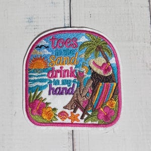 May include: A rectangular embroidered patch with rounded corners featuring a beach scene, a palm tree, a person in a lounge chair, and the text "toes in the sand drink in my hand" in pink, yellow, and blue thread on a white background.