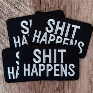 May include: Three rectangular embroidered patches with a black background and white stitched text that reads SHIT HAPPENS. Each patch features a clean, bold sans-serif font and a black merrowed border, arranged in a slightly overlapping stack.