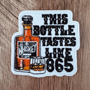 May include: An embroidered patch featuring an amber-colored whiskey bottle and a glass filled with liquid and ice. The text reads, "THIS BOTTLE TASTES LIKE 865" in bold black lettering. The patch has a white border and a white background.