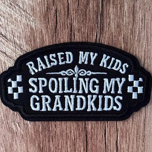 May include: A black embroidered patch with a scalloped border featuring white text that reads RAISED MY KIDS SPOILING MY GRANDKIDS. The design includes a central decorative flourish and a small chequered flag pattern on both the left and right sides.