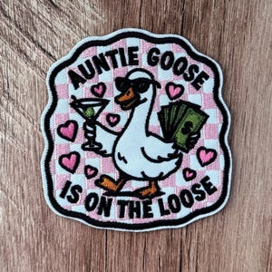 Könnte beinhalten: Ein bestickter Aufnäher mit einer weißen Gans, die eine schwarze Sonnenbrille trägt und ein Martiniglas sowie einen Fächer aus grünen Geldscheinen hält. Der Hintergrund ist ein rosa-weißes Karomuster mit rosa Herzen und dem Text AUNTIE GOOSE IS ON THE LOOSE.