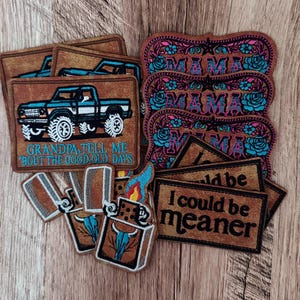 May include: A collection of rectangular and shaped embroidered patches featuring various designs. One patch shows a blue pickup truck with the text "GRANDPA, TELL ME 'BOUT THE GOOD OLD DAYS". Others feature the word "MAMA" with floral accents, lighters with bull skulls, and the text "I could be meaner".