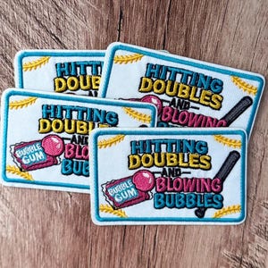 Peut inclure: Trois écussons rectangulaires brodés sur fond blanc avec une bordure bleue. Le texte HITTING DOUBLES AND BLOWING BUBBLES est écrit en bleu, jaune et rose. Chaque écusson présente un chewing-gum rose et une batte de baseball noire.
