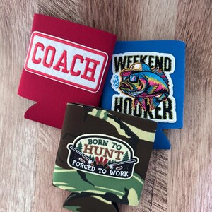 May include: Three foam can coolers featuring different designs. One red cooler displays the word COACH in white letters. One blue cooler features a colourful fish graphic with the text WEEKEND HOOKER. One camouflage cooler shows a hunting graphic with the text BORN TO HUNT FORCED TO WORK.