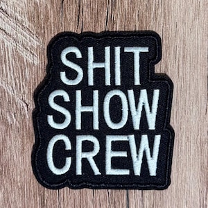 Puede incluir: Un parche bordado rectangular con fondo negro y letras blancas cosidas que dicen SHIT SHOW CREW. El parche presenta un borde negro grueso y una superficie de tela texturizada, con el texto dispuesto en tres líneas distintas.
