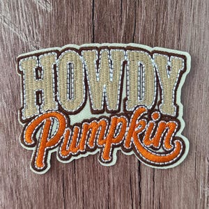 Puede incluir: Un parche bordado que presenta la palabra HOWDY en letras de bloque beige con contornos marrón oscuro, situada sobre la palabra Pumpkin en letra cursiva naranja con contornos marrón oscuro, sobre una base de tela color crema.