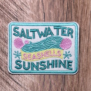 May include: A rectangular embroidered patch with a white background and a teal border. It features teal text reading SALTWATER, SEASHELLS, and SUNSHINE. The design includes teal embroidered waves, two pink seashells, and small teal starfish accents.