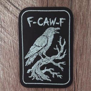 May include: A rectangular embroidered patch with a black background and a light grey border. The design features a light grey raven perched on a bare, gnarled tree branch. Above the bird, the text F-CAW-F is stitched in light grey thread.
