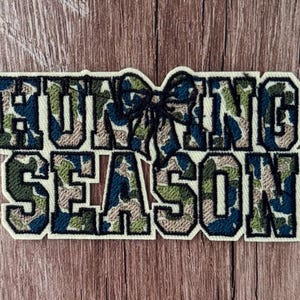 May include: An embroidered patch featuring the text HUNTING SEASON in a block font. The letters are filled with a camouflage pattern in shades of green, tan, and dark blue, with a black stitched bow icon replacing the letter T in HUNTING.