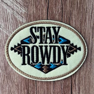 Könnte beinhalten: Ein ovaler, bestickter Aufnäher mit cremefarbenem Hintergrund und einem braunen Rand in Seiloptik. Der Text STAY ROWDY ist in schwarzem Garn gestickt, zentriert und von geometrischen Mustern in Schwarz, Braun und leuchtendem Blau umgeben.