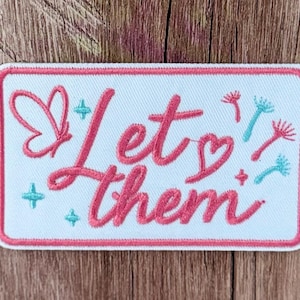 May include: A rectangular embroidered patch with a white background and a pink border. The text Let them is stitched in pink cursive. The design includes a pink butterfly outline, a small pink heart, and several teal and pink dandelion seeds.