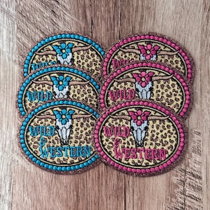 May include: Six oval embroidered patches featuring a white longhorn skull with a flower on its forehead, set against a tan and brown leopard print background. Three patches have blue borders and text, while three have pink borders and text reading WILD & WESTERN.