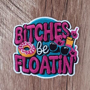 May include: An embroidered patch with a teal background featuring the text BITCHES be FLOATIN in bold pink and white lettering. The design includes a pink frosted donut with yellow sprinkles, black sunglasses, and a yellow drink with a pink straw.