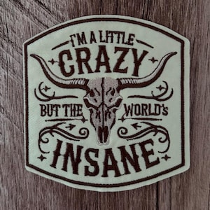 May include: An embroidered patch with a cream-colored background and dark brown stitching. The design features a central steer skull with long horns, surrounded by the text "I'M A LITTLE CRAZY BUT THE WORLD'S INSANE" in a decorative font.