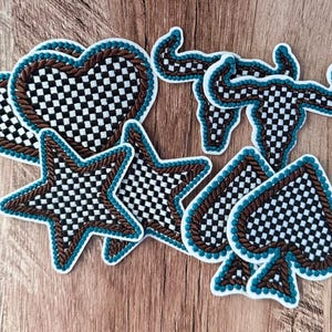 May include: A collection of embroidered patches featuring a black and white checkered pattern. The shapes include hearts, stars, spade symbols, and longhorn skulls. Each patch is outlined with a brown rope-like border and a thin teal edge.