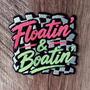 May include: A square embroidered patch featuring a black and white checkered background. The text "Floatin' & Boatin'" is stitched in bright pink and light green thread, with the word "Floatin'" in pink and "Boatin'" in green.