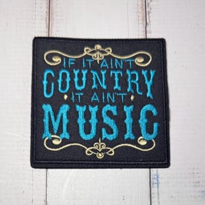 Puede incluir: Un parche cuadrado bordado en negro con el texto IF IT AIN'T COUNTRY IT AIN'T MUSIC en hilo azul brillante. El diseño está enmarcado por bordes decorativos de volutas de color dorado en la parte superior e inferior de las letras.