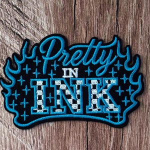 May include: An embroidered patch with a black background featuring the text Pretty in Ink in bright blue script and block lettering. The word INK has a white and blue checkered pattern. The design is surrounded by blue flame-like borders and stars.