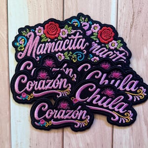 May include: A collection of black embroidered patches featuring floral designs and the words Mamacita, Corazón, and Chula in pink and purple thread. The patches have scalloped edges and include red roses, small green leaves, and decorative swirls.