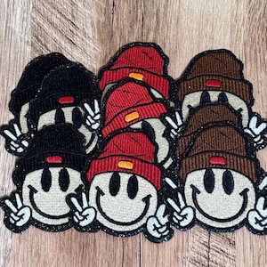 May include: A collection of embroidered patches featuring smiley faces wearing beanies in black, red, and brown. Each smiley face has black oval eyes, a wide grin, and hands making a peace sign. The beanies have a small rectangular accent.