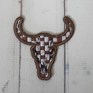 May include: An embroidered patch in the shape of a bull skull featuring a brown and white checkered pattern. The design includes a glittery brown border and dark stitched details around the eye sockets and nasal cavity of the skull shape.