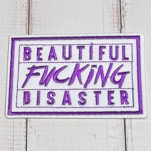 Puede incluir: Un parche rectangular bordado con fondo blanco y borde morado. El texto "BEAUTIFUL" está bordado en morado en la parte superior, "FUCKING" en una letra cursiva morada más grande en el medio y "DISASTER" en morado en la parte inferior.