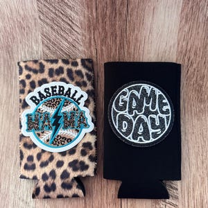 May include: Two foam can coolers. The left one features a brown and black leopard print with a circular patch displaying a baseball, a teal lightning bolt, and the text BASEBALL MAMA. The right one is solid black with a circular patch reading GAME DAY.