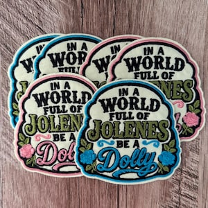 May include: A collection of embroidered fabric patches featuring an off-white background with black, olive green, pink, and blue thread. The text reads IN A WORLD FULL OF JOLENES BE A DOLLY, accented with embroidered pink and blue floral motifs.