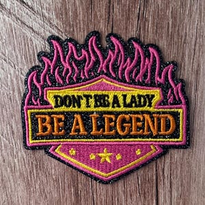 May include: A shield-shaped embroidered patch with a black glitter background and pink flame border. It features the yellow text DON'T BE A LADY and orange text BE A LEGEND. The lower section is pink with a yellow star and small yellow dots.