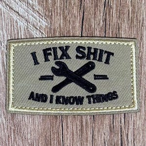 May include: A rectangular tan morale patch with a stitched border. The patch features black embroidered text that reads I FIX SHIT AND I KNOW THINGS, centered around a graphic of two crossed wrenches. The design is clean and clearly legible.