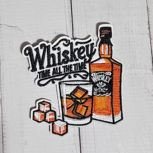 May include: An embroidered patch featuring a whiskey bottle, a glass filled with amber liquid and ice cubes, and three separate ice cubes. The text reads Whiskey TIME ALL THE TIME in black script and block lettering against a white background.