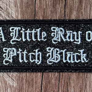May include: A rectangular black patch with a glittery finish featuring the white embroidered text A Little Ray of Pitch Black in a gothic font style. The patch has a thick black stitched border and is designed for attachment to fabric items.