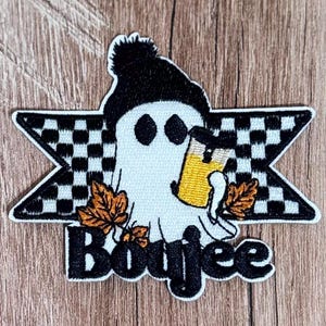 May include: An embroidered patch featuring a white ghost wearing a black beanie and holding a yellow drink. The ghost is centered over a black and white checkered star shape with orange autumn leaves at the base and the word Boujee in black.