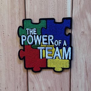 Peut inclure: Un écusson brodé de forme carrée composé de quatre pièces de puzzle imbriquées en vert, bleu, rouge et jaune. L'écusson présente une bordure noire épaisse et le texte brodé en blanc THE POWER OF A TEAM sur les pièces du puzzle.