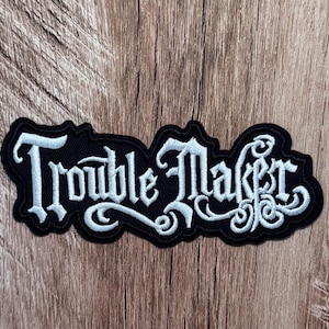 May include: A black embroidered patch featuring the text "Trouble Maker" in a stylized, white gothic-inspired font. The lettering has intricate, swirling flourishes at the ends of the characters, and the patch has a die-cut border following the shape.