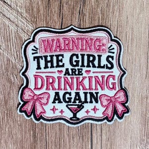 Op de afbeelding: Een geborduurde patch met een witte achtergrond en zwarte rand, voorzien van de tekst WARNING: THE GIRLS ARE DRINKING AGAIN in roze en zwarte letters, geaccentueerd met twee roze strikken, kleine roze hartjes en een martini-glas icoon.