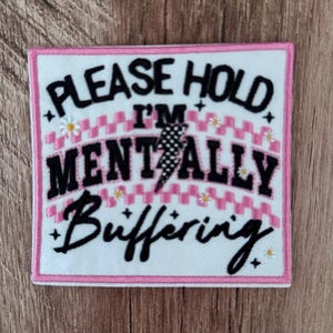 Peut inclure: Un écusson brodé carré à fond blanc avec une bordure rose. Le texte "PLEASE HOLD I'M MENTALLY BUFFERING" est écrit en noir, accompagné d'un éclair à pois noirs et blancs, de petites fleurs de marguerites blanches et d'étoiles noires.