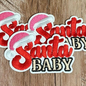 May include: Three embroidered iron-on patches featuring the text Santa BABY in red and gold thread. Each patch includes a pink and white Santa hat icon positioned above the word Santa. The patches have a white border and white backing.