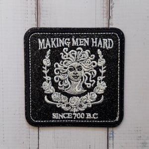 Op de afbeelding: Een vierkante zwarte glitterstoffen patch met witte borduursels van een Medusa-hoofd omringd door bloemenkransen. De tekst "MAKING MEN HARD" is bovenaan geborduurd en "SINCE 700 B.C" is onderaan geborduurd met witte draad.
