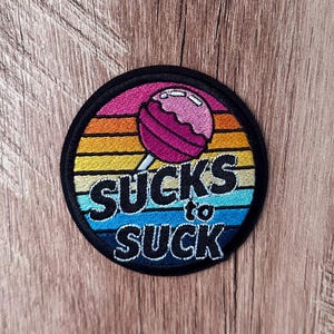 Puede incluir: Un parche bordado circular con un fondo de atardecer de estilo retro con rayas horizontales en rosa, naranja, amarillo y azul. Una piruleta rosa está centrada sobre el texto bordado en negro que dice SUCKS to SUCK.