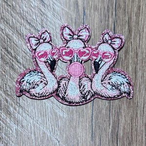 May include: An embroidered patch featuring three pink flamingos with glittery pink borders. Each flamingo wears a pink bow on its head and heart-shaped pink sunglasses. The central flamingo is blowing a pink bubble gum bubble.