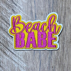 May include: An embroidered patch featuring the text Beach Babe in a stylized font. The word Beach is in magenta thread with a yellow outline, while the word Babe is in yellow thread with a magenta outline, all on a white fabric backing.