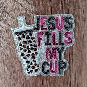 Puede incluir: Un parche de lentejuelas que muestra un vaso con un estampado de leopardo marrón y negro. El parche incluye el texto JESUS FILLS MY CUP en lentejuelas negras y rosas sobre un fondo de purpurina plateada, creando un accesorio decorativo.