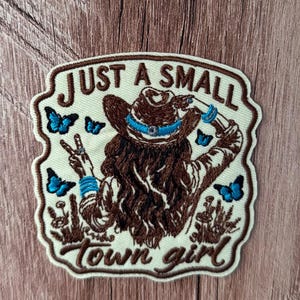 May include: An embroidered patch on a cream background featuring a person with long brown hair wearing a brown cowboy hat with a blue band. The person makes a peace sign. Text reads JUST A SMALL town girl. Blue butterflies and wildflowers surround the figure.
