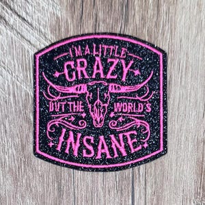 May include: A black glittery embroidered patch with a pink border and pink text that reads I'M A LITTLE CRAZY BUT THE WORLD'S INSANE. The design features a central pink steer skull graphic with long horns and decorative scrollwork accents.
