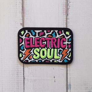 May include: A rectangular embroidered patch with a black border featuring a white background with a multi-colored leopard print pattern. The text ELECTRIC SOUL is stitched in pink and lime green, flanked by two orange lightning bolt icons.
