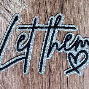 May include: A decorative patch featuring the black embroidered text Let Them in a cursive script font. The lettering is outlined with a thick, shimmering silver glitter border, and a small black embroidered heart is positioned at the end.