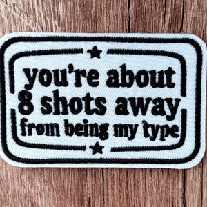 May include: A rectangular embroidered patch with a white background and a black border. The text reads, "you're about 8 shots away from being my type," in black lettering, with a small black star centered above and below the text.